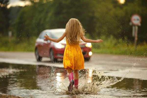 A little girl in a yellow dress runs through the puddles and spl — Stock Photo, Image