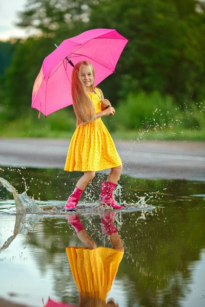 Een klein meisje in een gele jurk met een roze paraplu loopt throug — Stockfoto
