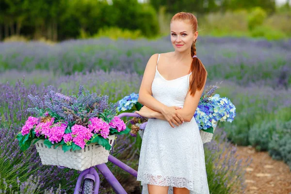 Menina de cabelos vermelhos com um pigtail em um vestido branco em uma bicicleta roxa — Fotografia de Stock