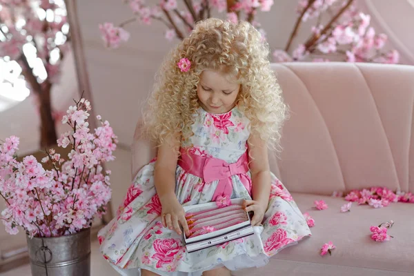 Маленькая очень милая кудрявая блондинка в платье сидит на диване и — стоковое фото