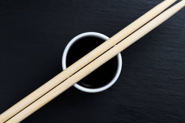 Soya sosu küçük beyaz kasede, yemek çubuklarıyla siyah mutfak tabağında. Tipik Asya mutfağı baharatı.