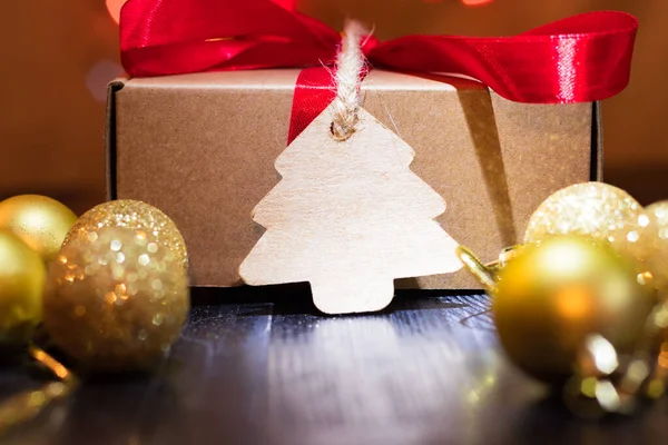 Christmas present with label with twine in the spotlight. Red ribbon bow. Black wooden table. Yellow tree baubles.