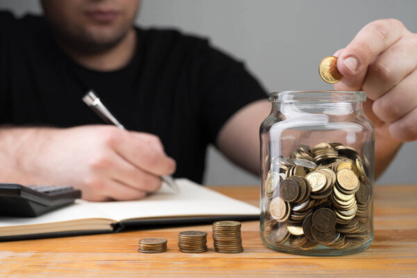 Coins in a jar, stacks of change and young caucasian man dropping money and writing in a notebook. Tax forms, counting savings, money collecting or accounting abstract concept.