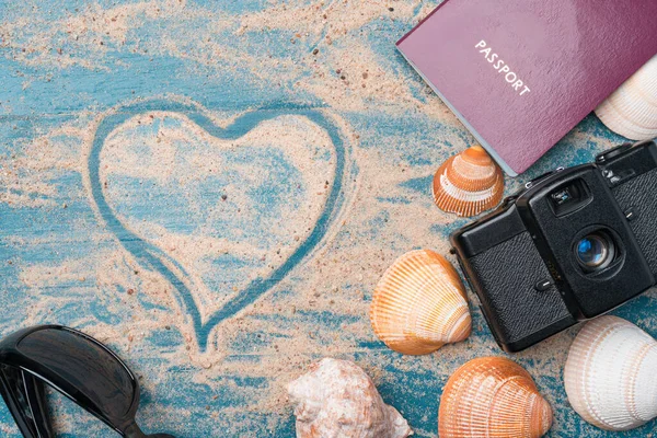 Passport, vintage film camera, sunglasses and seashells on a blue wooden background with golden beach sand and heart shape. Summer travel love abstract concept. Copy space on the left.