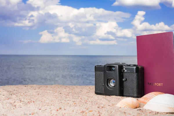 Passport, vintage film camera and seashells on golden beach sand. Summer travel abstract concept. Copy space on the left.