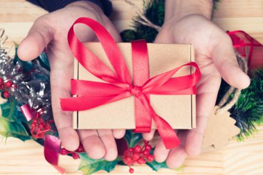 Elinde hediye ya da hediye tutan zarif bir adam. Kırmızı kurdele. Noel süsleri. Doğal ahşap masa. Düz manzaralı. Eski renkler.