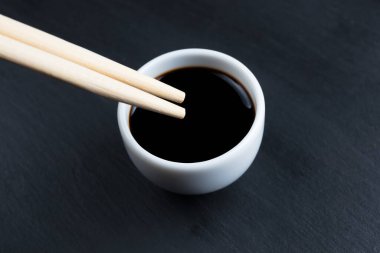 Soya sosu küçük beyaz kasede, yemek çubuklarıyla siyah mutfak tabağında. Tipik Asya mutfağı baharatı.