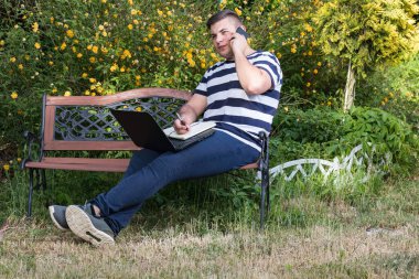 Genç bir iş adamı ya da iş adamı. Laptop ve akıllı telefonla bahçede çalışıyor. Ofis özgürlüğü dışında serbest.