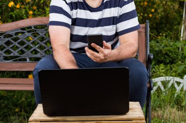 Genç bir iş adamı ya da iş adamı. Laptop ve akıllı telefonla bahçede çalışıyor. Ofis özgürlüğü dışında serbest.