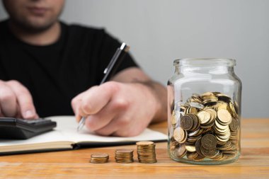 Kavanozdaki madeni paralar, bozuk paralar ve vergi formlarını dolduran genç beyaz adam ya da arka plandaki hesap makinesindeki tasarrufları saymak. Para soyut kavramları topluyor. Boşluğu kopyala.