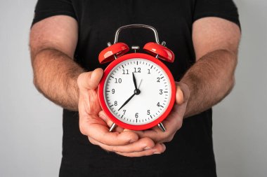 Siyah tişörtlü beyaz adam elinde klasik kırmızı alarmlı saat tutuyor.