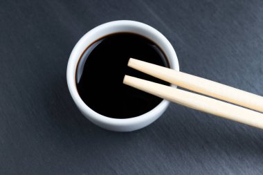 Soya sosu küçük beyaz kasede, yemek çubuklarıyla siyah mutfak tabağı arka planında. Tipik Asya mutfağı baharatı.