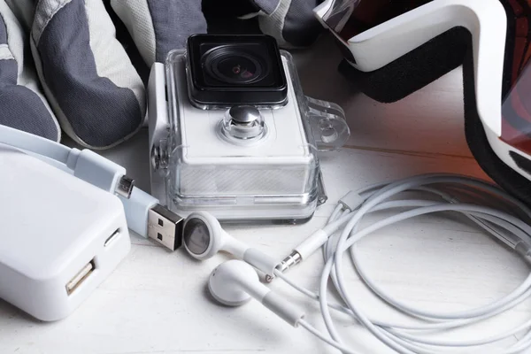 White technology on natural wooden table background. Power bank, action camera, head phones, cable goggles, gloves. Active winter leisure time sports holidays abstract concept. Skiing, snowboarding.