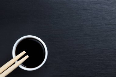 Soya sosu küçük beyaz kasede, yemek çubuklarıyla siyah mutfak tabağında. Tipik Asya mutfağı baharatı. Boşluğu sağda kopyala.