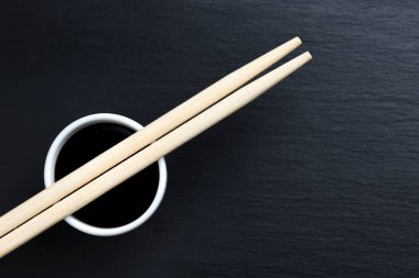 Soya sosu küçük beyaz kasede, yemek çubuklarıyla siyah mutfak tabağında. Tipik Asya mutfağı baharatı. Boşluğu sağda kopyala.