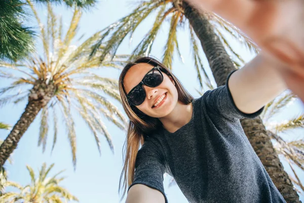 Joven mujer hermosa turista o blogger hace una selfie o está informando a sus suscriptores en el fondo de las palmeras en un país caliente — Foto de Stock
