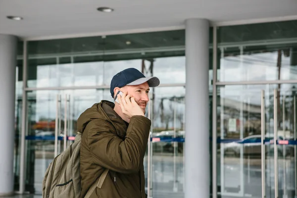 Молодой турист в аэропорту или рядом с торговым центром или вокзалом вызывает такси, разговаривает по мобильному телефону или общается с друзьями с помощью мобильного телефона — стоковое фото