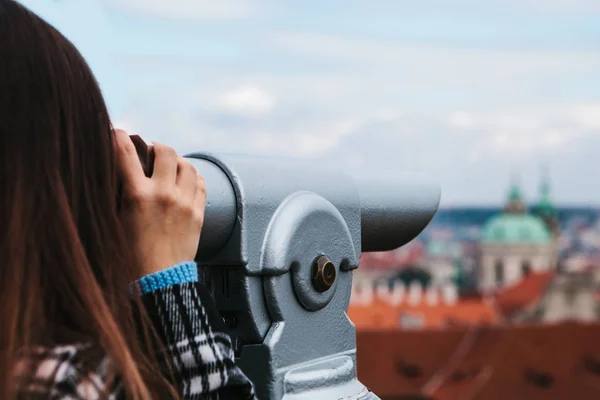 Девушка смотрит через бинокль на красивую архитектуру Праги на смотровой площадке. Прага является одним из самых любимых мест для посещения туристов — стоковое фото