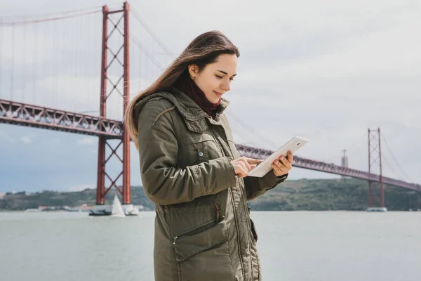 Una mujer joven y hermosa utiliza una tableta para comunicarse con amigos o mira un mapa o algo más. Puente 25 de abril en Lisboa en Portugal en el fondo . — Foto de Stock