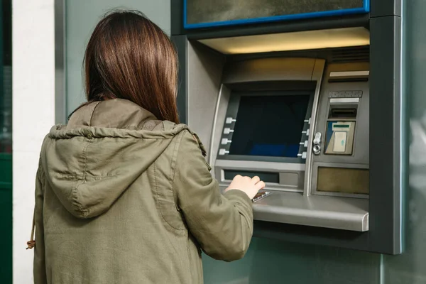 Una mujer joven toma dinero de un cajero automático. Toma una tarjeta del cajero automático. Finanzas, tarjeta de crédito, retiro de dinero . — Foto de Stock