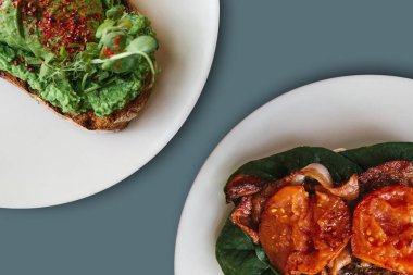 Yakın çekim. Tabaklarda bir sandviç ile avokado ve yanında bir sandviç ya da tost pastırma ve minimalist tarzda kızarmış domates ile oldu. Sağlıklı ve sağlıksız gıdalar arasında seçme kavramı.