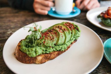Sandviç ya da tost ile avokado ön planda. Bir kişi ve içecek kahve veya çay yemek olacak
