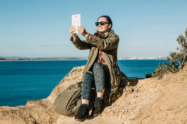 Una joven hermosa chica hace selfie o habla mediante el uso de una tableta en línea o toma fotos de un hermoso paisaje — Foto de Stock