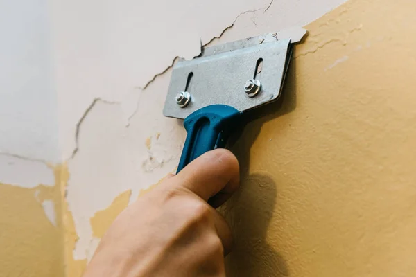 Un hombre con un rascador quita la vieja cubierta de la pared . — Foto de Stock