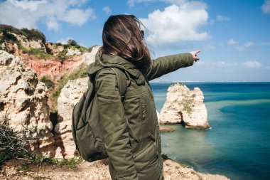 Genç kadın turist Atlantik Okyanusu ve Portekiz kıyılarında manzara güzel manzaralarına sahip bir sırt çantası ile bir el mesafe gösterir.