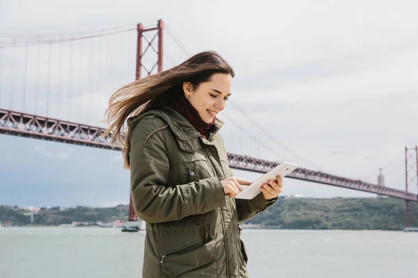 Una mujer joven y hermosa utiliza una tableta para comunicarse con amigos o mira un mapa o algo más. Puente 25 de abril en Lisboa en Portugal en el fondo . — Foto de Stock