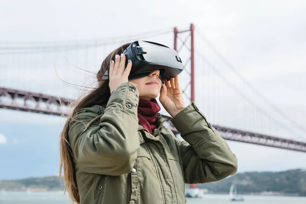 Joven hermosa chica con gafas de realidad virtual. Puente del 25 de abril en Lisboa en el fondo. El concepto de las tecnologías modernas y su uso en la vida cotidiana —  Fotos de Stock