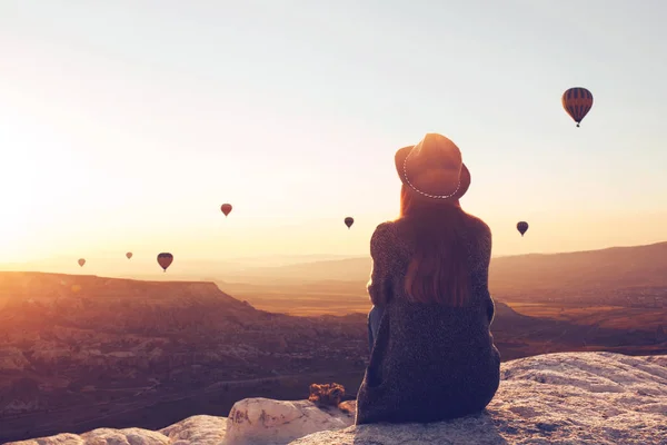View from the back of a girl in a hat sits on a hill and looks at air balloons. — Stock Photo, Image