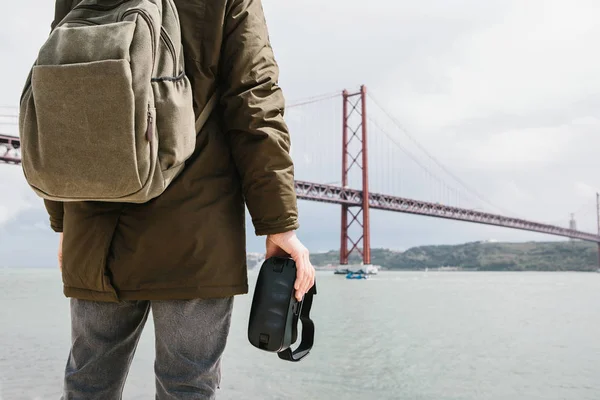 The man holds the virtual reality glasses and looks at the 25th of April bridge in Lisbon. The concept of virtual travel and tourism. Modern technologies and their use in everyday life