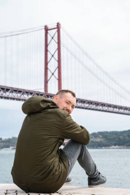 Genç bir adam Lizbon sahildeki oturur ve kameraya benziyor.
