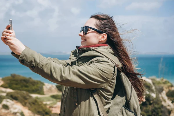 Una chica turística positiva toma una hermosa vista o hace una selfie o se comunica con alguien en línea utilizando un teléfono celular — Foto de Stock