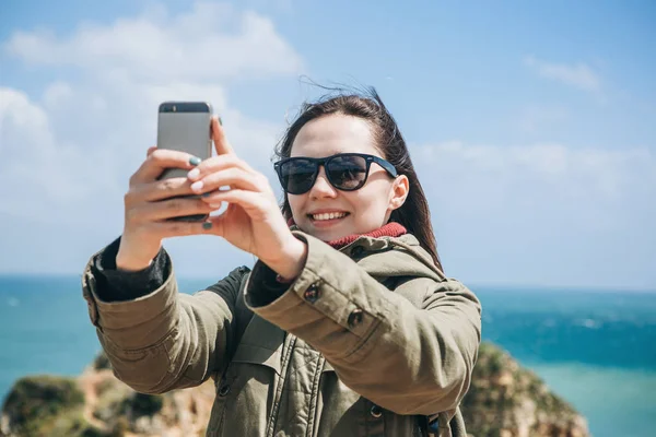 Una chica turística positiva toma una hermosa vista o hace una selfie o se comunica con alguien en línea utilizando un teléfono celular — Foto de Stock