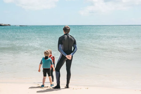 Mladý surfař se svými dětmi na pláži se dívá na moři nebo oceánu. Společná zábava. Šťastná rodina — Stock fotografie