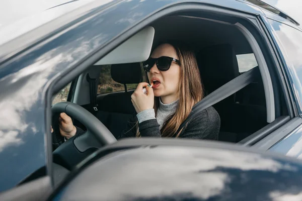 Linda chica joven conductor pinta los labios dentro del coche — Foto de Stock