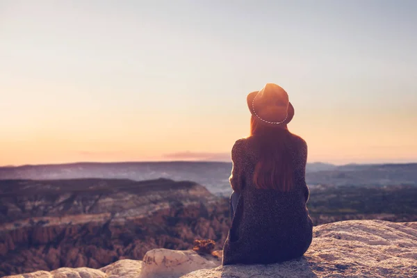 A view from the back of a girl in a hat sits on a hill and admires the beautiful view. — Stock Photo, Image