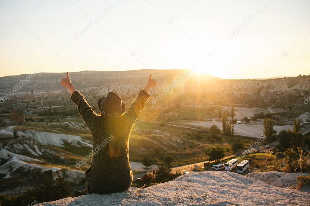 A tourist girl in a hat sits on a mountain and looks at the sunrise.