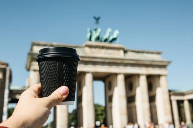 Bir kişi tek kullanımlık bir fincan kahve ya da Berlin Brandenburg Kapısı'nın arka planda başka bir içecek ile tutar.