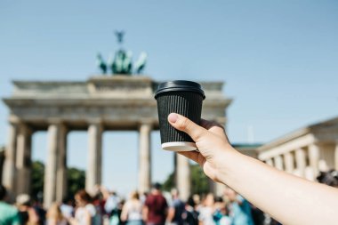 Bir kişi tek kullanımlık bir fincan kahve ya da Berlin Brandenburg Kapısı'nın arka planda başka bir içecek ile tutar.