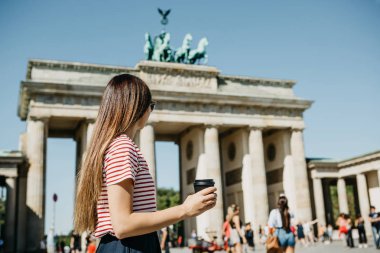 Bir kişi tek kullanımlık bir fincan kahve ya da Berlin Brandenburg Kapısı'nın arka planda başka bir içecek ile tutar.