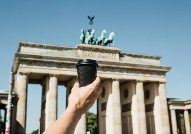 Bir kişi tek kullanımlık bir fincan kahve ya da Berlin Brandenburg Kapısı'nın arka planda başka bir içecek ile tutar.