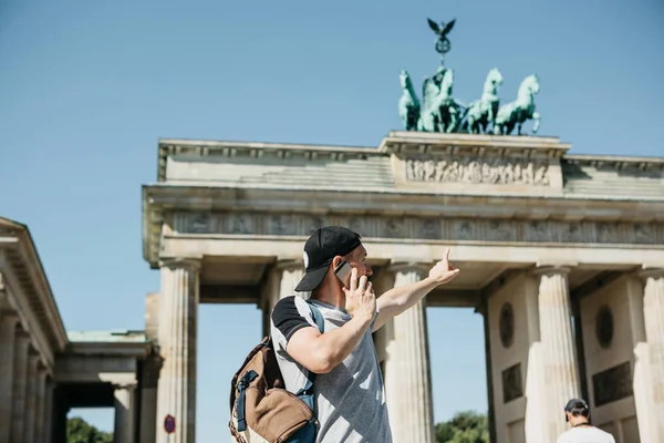 Jako turista nebo chlap s batohem mluví na mobilním telefonu a ukazuje rukou na Braniborská brána v Berlíně — Stock fotografie