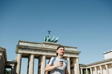 Bir kişi tek kullanımlık bir fincan kahve ya da Berlin Brandenburg Kapısı'nın arka planda başka bir içecek ile tutar.