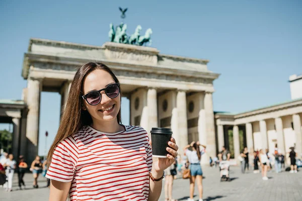 Eine person hält eine einwegbecher mit kaffee oder einem anderen getränk auf dem hintergrund des brandenburger tores in berlin. — Stockfoto