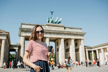 Bir kişi tek kullanımlık bir fincan kahve ya da Berlin Brandenburg Kapısı'nın arka planda başka bir içecek ile tutar.