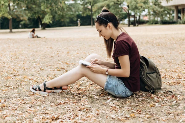Uma jovem com uma mochila senta-se no parque de outono e usa o tablet . — Fotografia de Stock