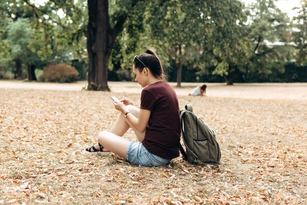 Дівчина з рюкзаком сидить в осінньому парку і використовує мобільний телефон . — стокове фото